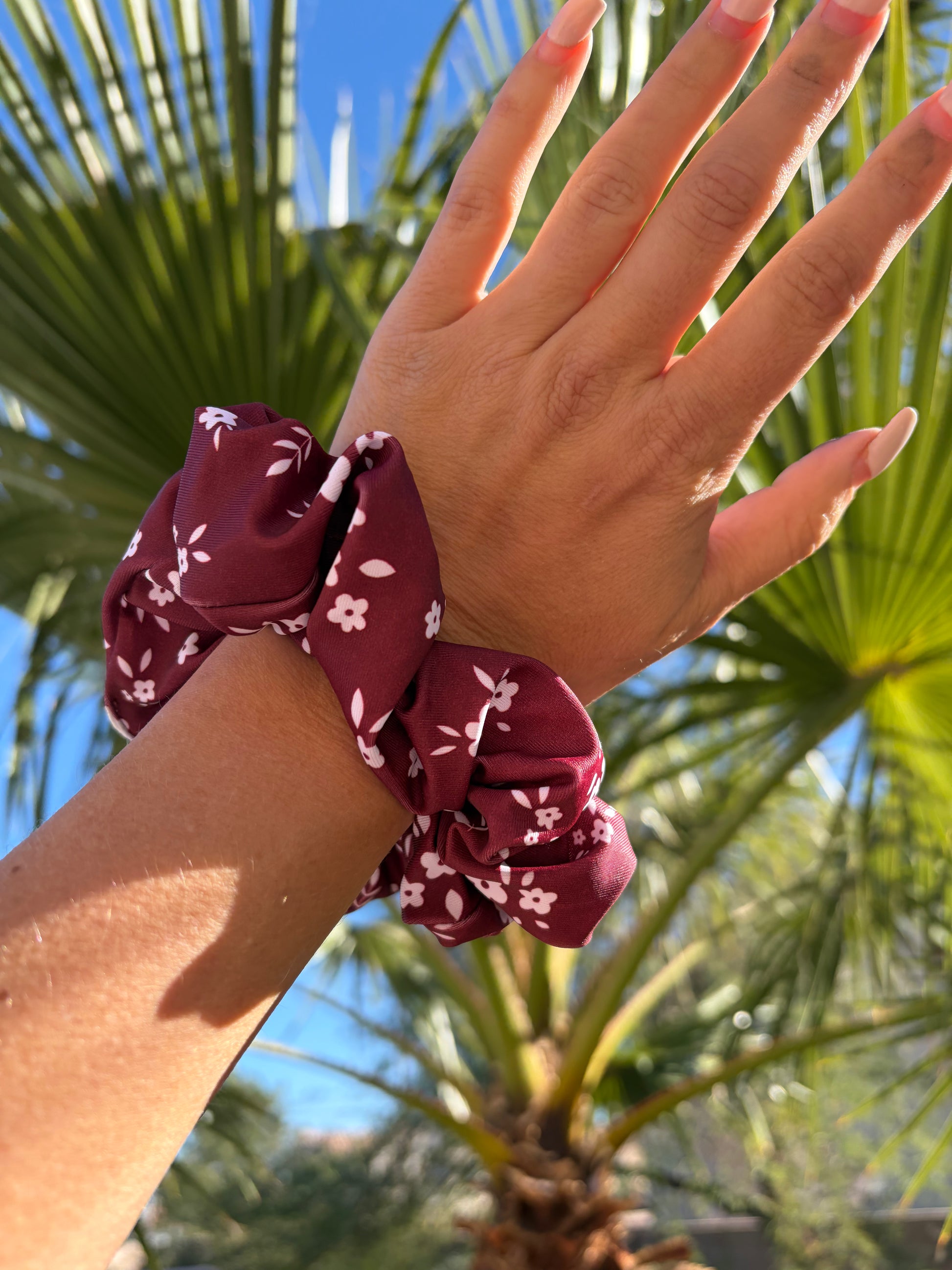 Hand wearing a maroon scrunchie with white floral pattern against a palm tree background