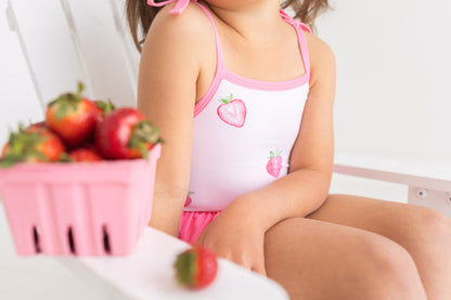 Child wearing a strawberry-themed swimsuit sitting next to a basket of strawberries.